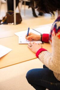 A woman writing notes in a notebook during a classroom lecture setting.